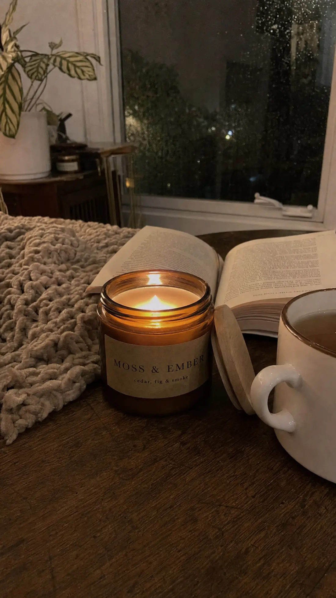 The candle lit beside an open book, a mug of tea and a knitted blanket