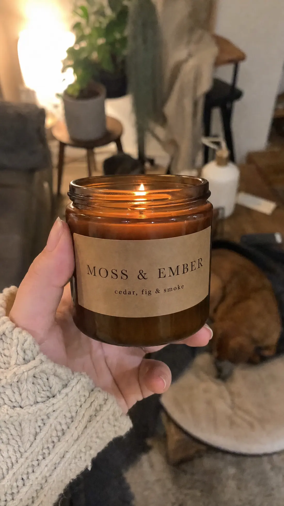 Someone holding up a lit Moss & Ember candle in a cosy living room